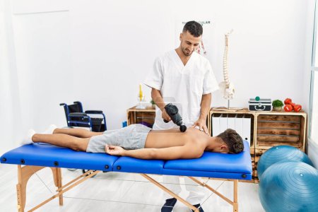 Téléchargez les photos : Deux hommes hispaniques physiothérapeute et patient massant le dos à l'aide d'un pistolet à percussion à la clinique - en image libre de droit