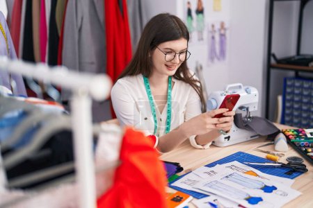 Téléchargez les photos : Jeune femme caucasienne tailleur souriant confiant en utilisant un smartphone au magasin de tailleurs - en image libre de droit