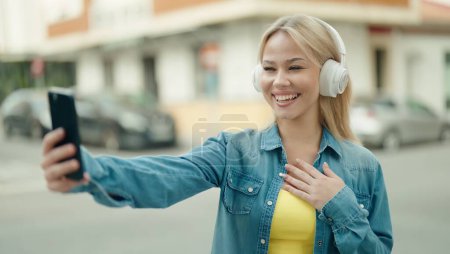 Téléchargez les photos : Jeune femme blonde souriante confiante ayant appel vidéo à la rue - en image libre de droit