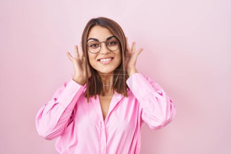 Téléchargez les photos : Young hispanic woman wearing glasses standing over pink background trying to hear both hands on ear gesture, curious for gossip. hearing problem, deaf - en image libre de droit