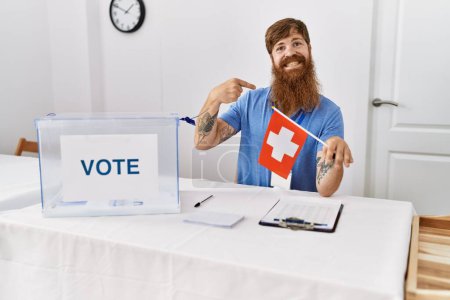 Foto de Hombre caucásico con barba larga en las elecciones de campaña política sosteniendo bandera suiza señalando con el dedo a uno mismo sonriendo feliz y orgulloso - Imagen libre de derechos