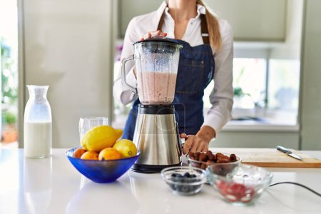 Photo for Young blonde woman liquefying smoothie using bender at kitchen - Royalty Free Image