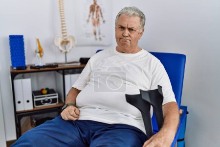 Foto de Hombre caucásico mayor en la clínica de fisioterapia sosteniendo muletas deprimido y la preocupación por la angustia, llorando enojado y con miedo. expresión triste. - Imagen libre de derechos