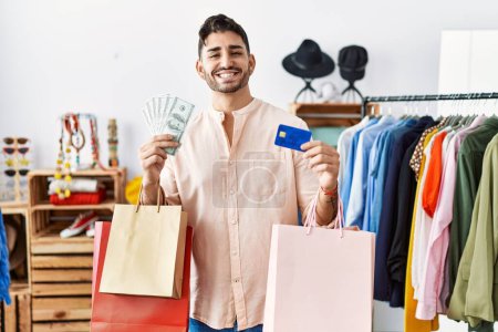 Foto de Joven hispano sosteniendo bolsas de compras y tarjeta de crédito sonriendo con una sonrisa alegre y fresca en la cara. mostrando dientes. - Imagen libre de derechos