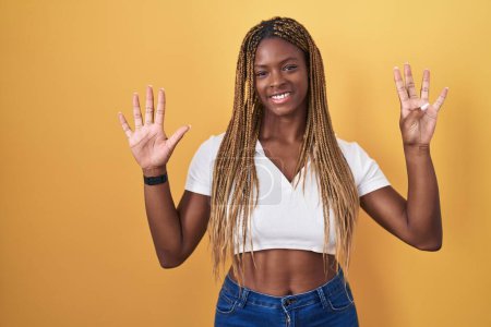 Foto de Mujer afroamericana con el pelo trenzado de pie sobre fondo amarillo mostrando y apuntando hacia arriba con los dedos número nueve mientras sonríe confiado y feliz. - Imagen libre de derechos