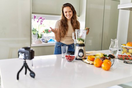 Photo for Young beautiful hispanic woman preparing vegetable smoothie with blender recording video tutorial with camera at the kitchen - Royalty Free Image