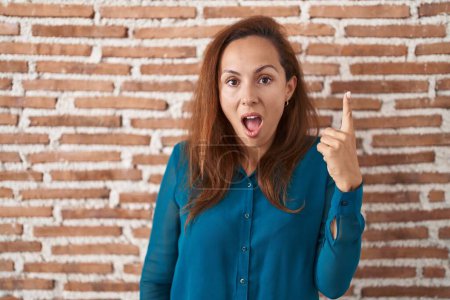 Téléchargez les photos : Femme brune debout sur un mur de briques pointant du doigt avec une idée réussie. Sorti et heureux. numéro un. - en image libre de droit