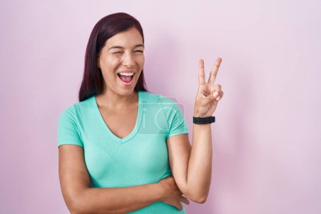 Foto de Mujer hispana joven de pie sobre fondo rosa sonriendo con la cara feliz guiñando el ojo a la cámara haciendo signo de victoria con los dedos. número dos. - Imagen libre de derechos