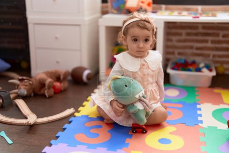 Téléchargez les photos : Adorable fille caucasienne jouer avec poupée assis sur le sol à la maternelle - en image libre de droit