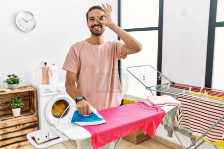 Foto de Joven hombre hispano planchando ropa en casa haciendo buen gesto con la mano sonriente, los ojos mirando a través de los dedos con cara feliz. - Imagen libre de derechos