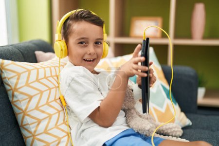 Foto de Adorable niño hispano usando touchpad y auriculares sentados en el sofá en casa - Imagen libre de derechos