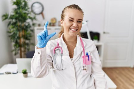 Téléchargez les photos : Young caucasian woman wearing doctor uniform and stethoscope at the clinic smiling and confident gesturing with hand doing small size sign with fingers looking and the camera. measure concept. - en image libre de droit