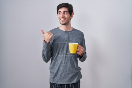 Téléchargez les photos : Young hispanic man wearing pajama drinking a cup of coffee smiling with happy face looking and pointing to the side with thumb up. - en image libre de droit