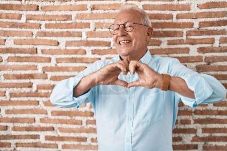 Photo for Senior man with grey hair standing over bricks wall smiling in love doing heart symbol shape with hands. romantic concept. - Royalty Free Image