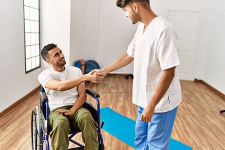 Téléchargez les photos : Deux hommes hispaniques physiothérapeute et patient assis en fauteuil roulant ayant séance de désintox serrent la main à la clinique - en image libre de droit
