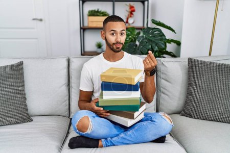 Téléchargez les photos : African american young man holding a pile of books sitting on the sofa doing money gesture with hands, asking for salary payment, millionaire business - en image libre de droit