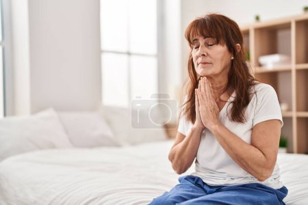 Foto de Middle age woman praying sitting on bed at bedroom - Imagen libre de derechos