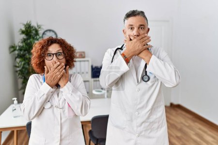 Foto de Two middle age doctors at medical clinic shocked covering mouth with hands for mistake. secret concept. - Imagen libre de derechos