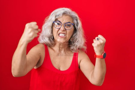 Téléchargez les photos : Femme d'âge moyen avec les cheveux gris debout sur fond rouge en colère et fou levant les poings frustrés et furieux tout en criant avec colère. rage et concept agressif. - en image libre de droit