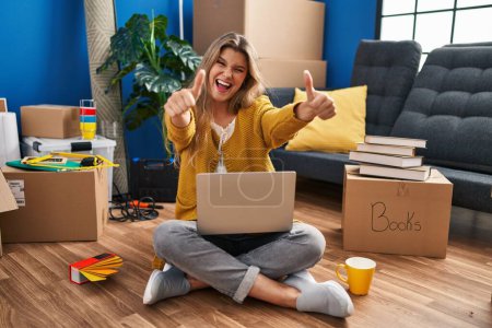 Photo for Young woman sitting on the floor at new home using laptop approving doing positive gesture with hand, thumbs up smiling and happy for success. winner gesture. - Royalty Free Image