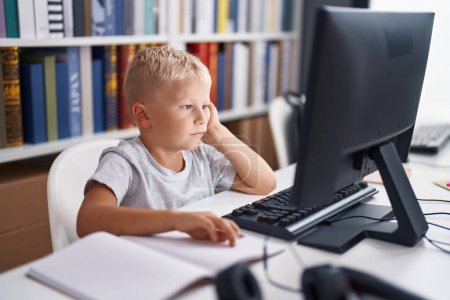 Foto de Adorable estudiante pequeño aburrido usando la computadora sentada en la mesa en el aula - Imagen libre de derechos
