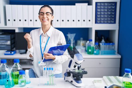 Téléchargez les photos : Jeune femme brune travaillant au laboratoire scientifique très heureuse et excitée de faire geste gagnant avec les bras levés, souriant et criant pour le succès. concept de célébration. - en image libre de droit