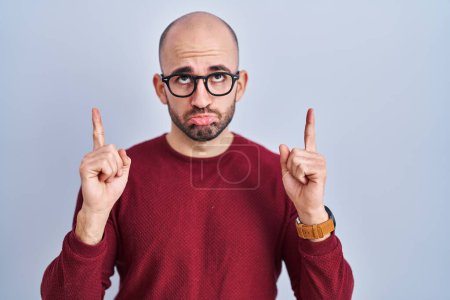 Téléchargez les photos : Jeune homme chauve avec la barbe debout sur fond blanc portant des lunettes pointant vers le haut l'air triste et bouleversé, indiquant la direction avec les doigts, malheureux et déprimé. - en image libre de droit