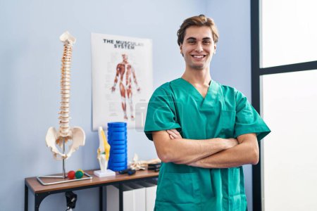 Téléchargez les photos : Jeune homme caucasien pysiothérapeute souriant confiant debout avec les bras croisés geste à la clinique de réadaptation - en image libre de droit