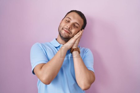 Foto de Hombre hispano de pie sobre fondo rosa durmiendo cansado soñando y posando con las manos juntas mientras sonríe con los ojos cerrados. - Imagen libre de derechos