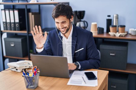 Foto de Hombre latino guapo trabajando en la oficina usando un ordenador portátil con aspecto positivo y feliz de pie y sonriendo con una sonrisa confiada mostrando los dientes - Imagen libre de derechos