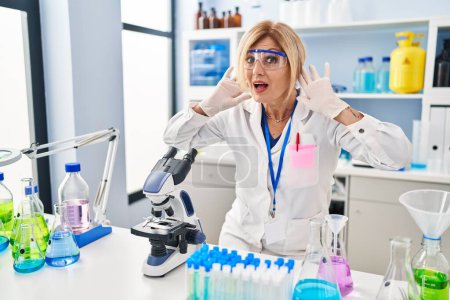 Photo for Middle age blonde woman working at scientist laboratory trying to hear both hands on ear gesture, curious for gossip. hearing problem, deaf - Royalty Free Image
