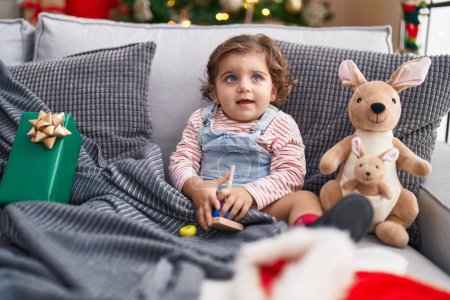 Foto de Adorable hispanic girl playing with hoops game sitting on sofa by christmas tree at home - Imagen libre de derechos