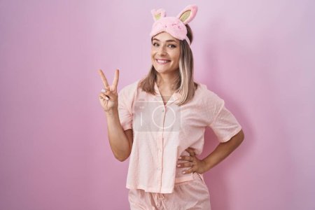 Téléchargez les photos : Blonde caucasian woman wearing sleep mask and pajama smiling with happy face winking at the camera doing victory sign with fingers. number two. - en image libre de droit