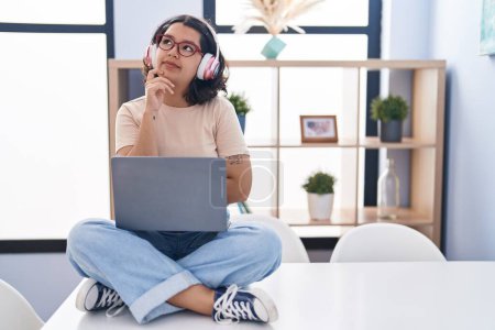 Photo for Young hispanic woman using laptop sitting on the table wearing headphones with hand on chin thinking about question, pensive expression. smiling with thoughtful face. doubt concept. - Royalty Free Image