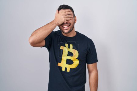 Photo for Young hispanic man wearing bitcoin t shirt smiling and laughing with hand on face covering eyes for surprise. blind concept. - Royalty Free Image