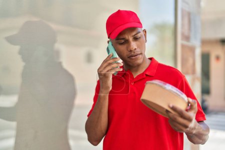 Photo for Young latin man delivery worker holding take away food talking on smartphone at street - Royalty Free Image