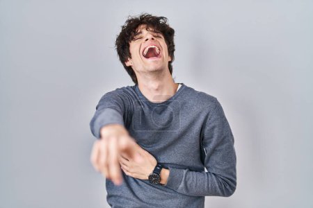 Photo for Young man standing over isolated background laughing at you, pointing finger to the camera with hand over body, shame expression - Royalty Free Image