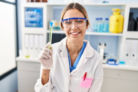 Foto de Mujer rubia joven que trabaja en el laboratorio científico sosteniendo la muestra con un aspecto positivo y feliz de pie y sonriendo con una sonrisa confiada mostrando los dientes - Imagen libre de derechos