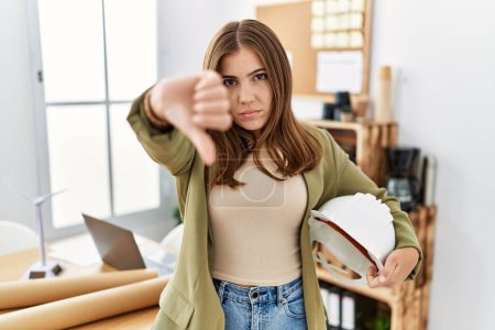 Photo for Young brunette woman holding architect hardhat at the office with angry face, negative sign showing dislike with thumbs down, rejection concept - Royalty Free Image