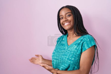 Photo for Young african american with braids standing over pink background inviting to enter smiling natural with open hand - Royalty Free Image