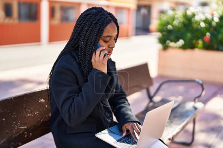Photo for African american woman using laptop talking on smartphone at street - Royalty Free Image