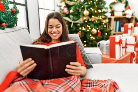 Photo for Young latin woman reading book sitting by christmas tree at home - Royalty Free Image