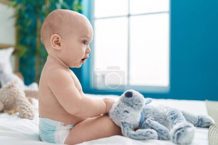 Téléchargez les photos : Adorable bébé caucasien assis sur le lit avec une expression détendue dans la chambre - en image libre de droit
