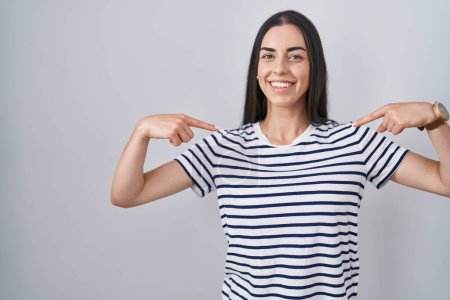 Téléchargez les photos : Jeune femme brune portant un t-shirt rayé à l'air confiant avec le sourire sur le visage, se montrant du doigt fier et heureux. - en image libre de droit