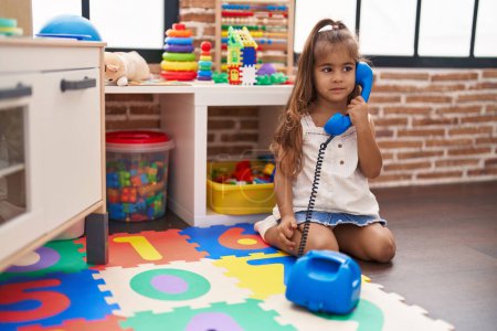 Foto de Adorable chica hispana jugando juguete telefónico sentado en el suelo en el jardín de infantes - Imagen libre de derechos