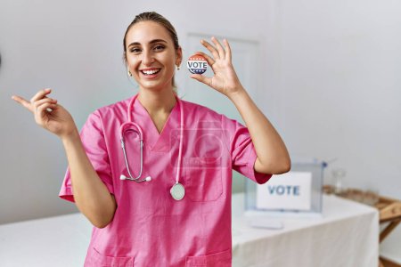 Téléchargez les photos : Young nurse woman at political campaign holding usa vote badge smiling happy pointing with hand and finger to the side - en image libre de droit