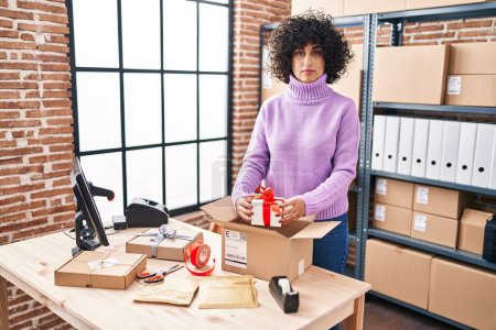 Téléchargez les photos : Young brunette woman with curly hair working at small business ecommerce preparing order thinking attitude and sober expression looking self confident - en image libre de droit