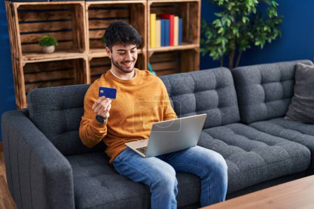 Photo for Young hispanic man using laptop and credit card sitting on sofa at home - Royalty Free Image