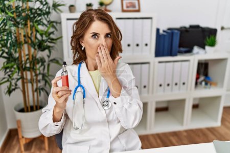 Photo for Middle age doctor woman holding electronic cigarette at medical clinic covering mouth with hand, shocked and afraid for mistake. surprised expression - Royalty Free Image