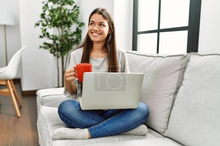 Téléchargez les photos : Jeune femme latine utilisant un ordinateur portable et boire du café assis sur le canapé à la maison - en image libre de droit